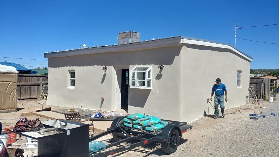 Small beige house under construction with a worker and a trailer in a dusty yard