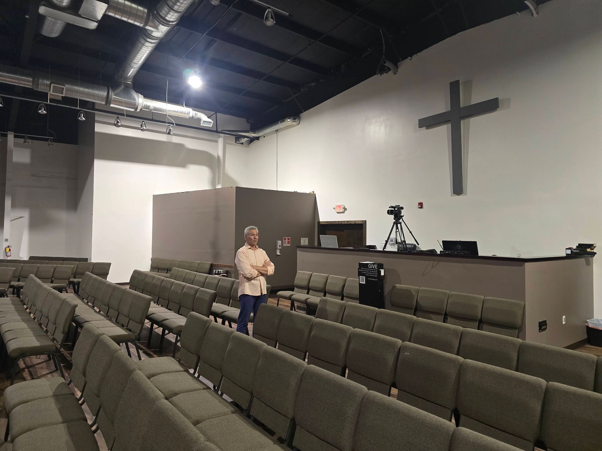 Empty auditorium with rows of gray chairs, a person standing near the front, and a large cross on the wall.
