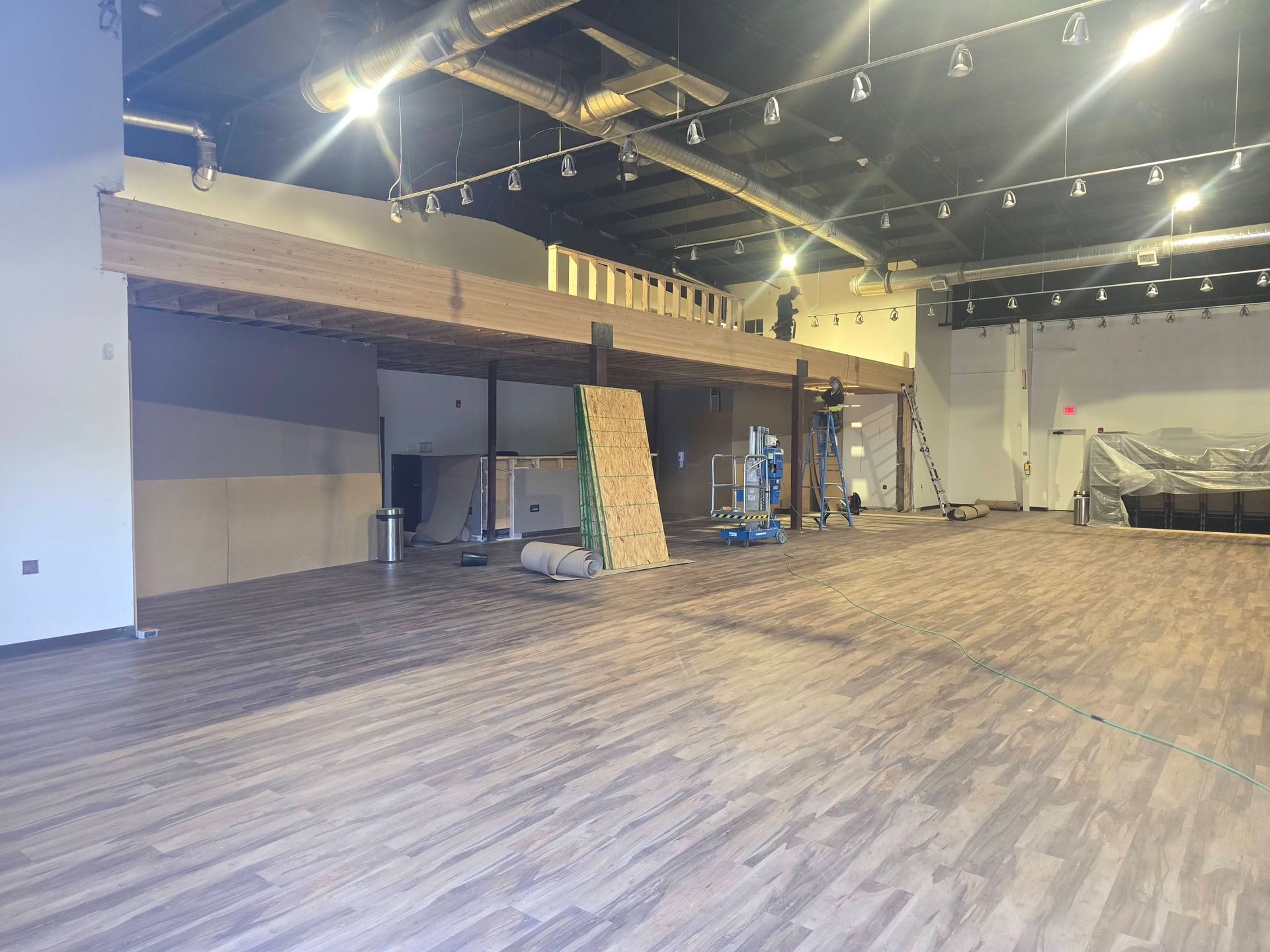 Empty indoor event hall with wood-pattern flooring, string lights, and a stage area on the right.