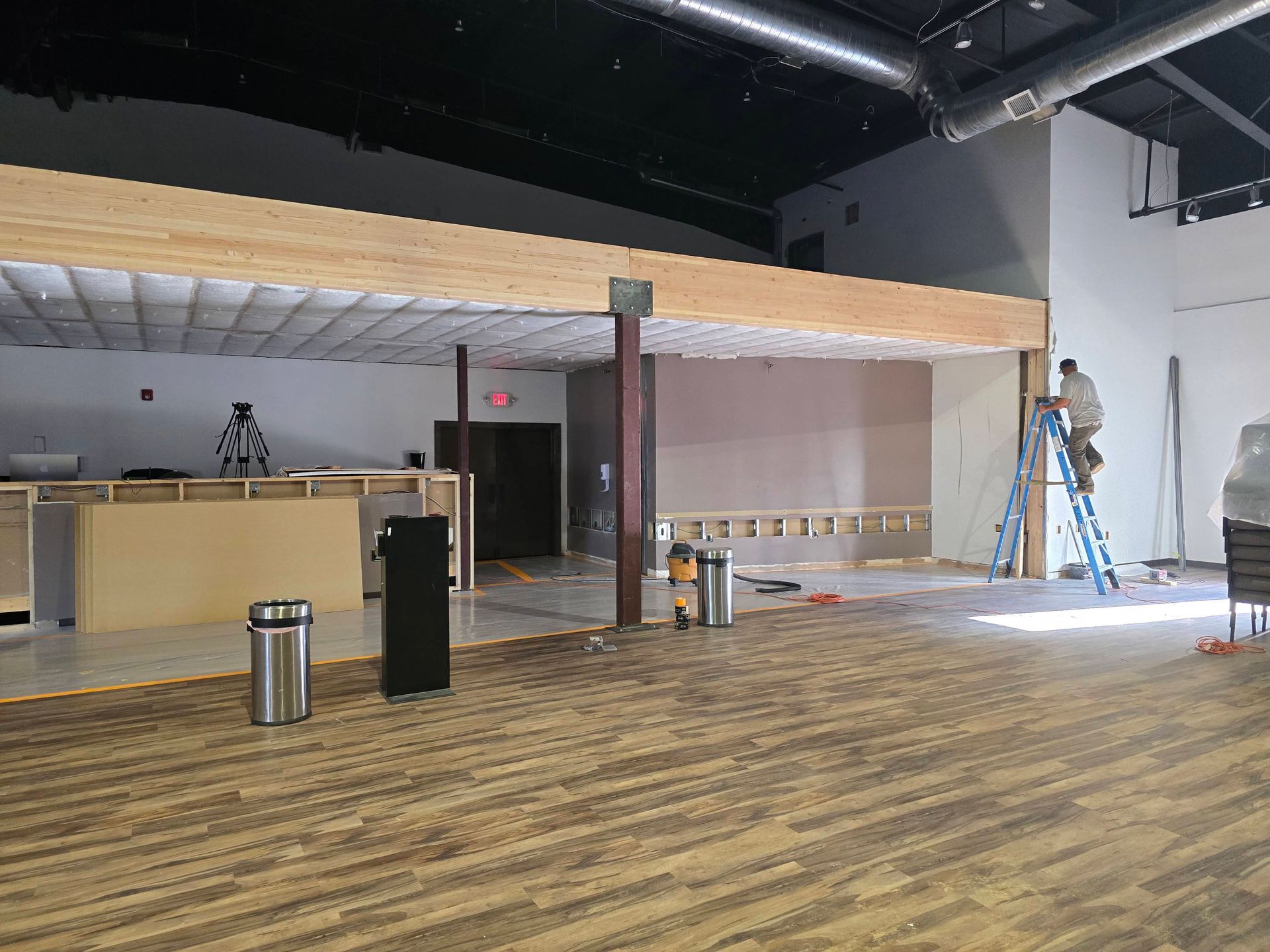 Empty indoor hall with wood floor, mezzanine railing, tables, trash cans, and a ladder by a white wall