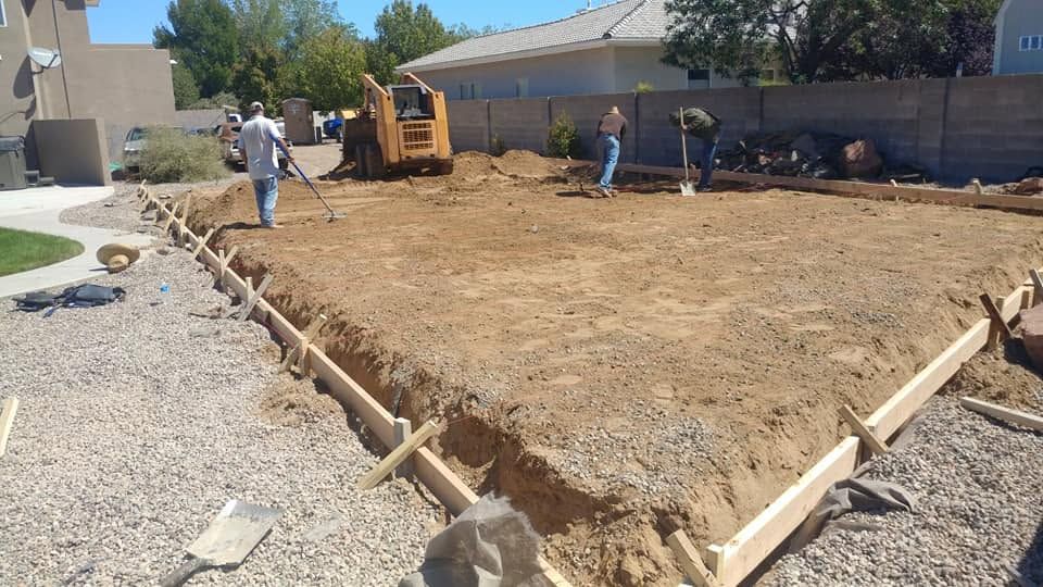 Construction crew grading a dirt lot with excavator and wooden formwork beside a gravel path