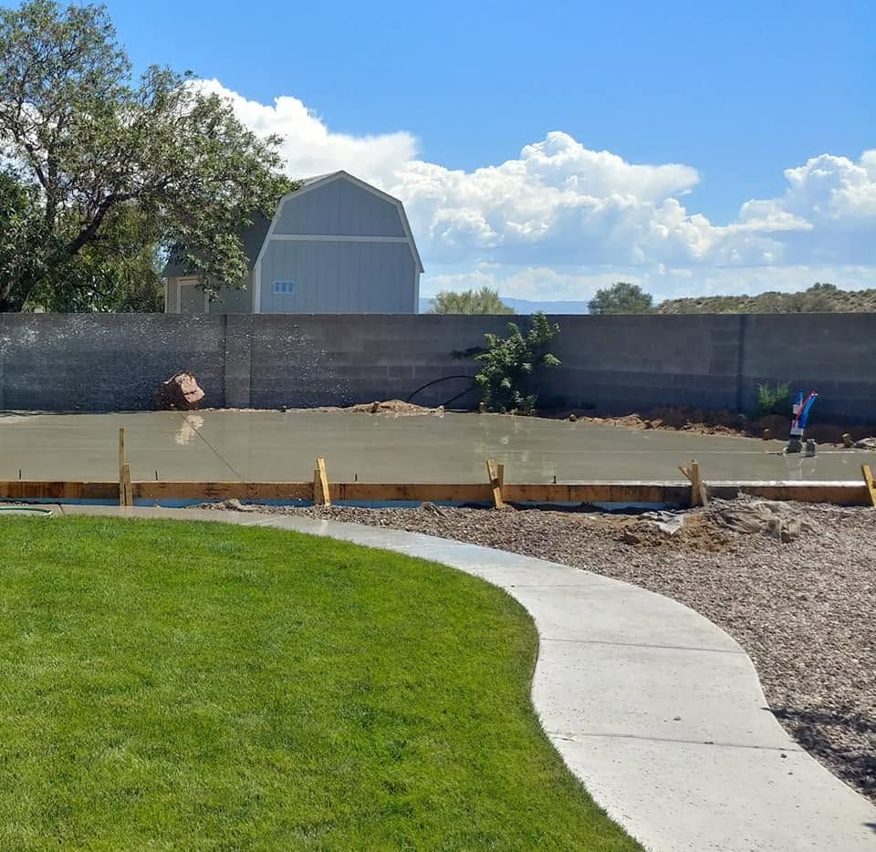 Backyard under construction with a concrete patio edge, curved walkway, grass, and a gray fence.