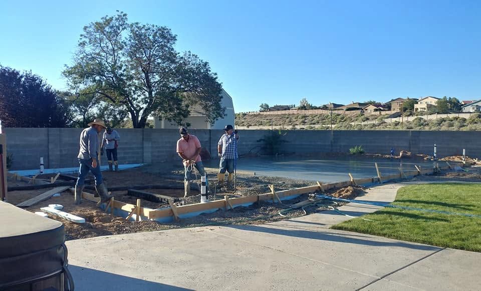 Workers pouring concrete for a backyard patio near a pool, with forms and tools in a sunny yard.