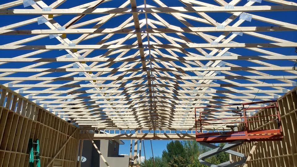 Wooden roof framing under construction against a blue sky, with a ladder and scaffolding visible below.