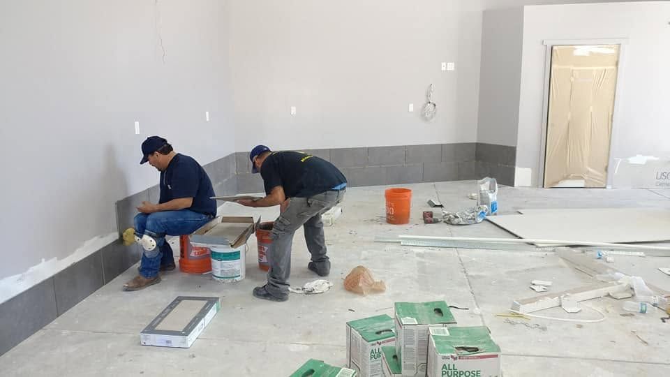 Two workers kneel on a tiled floor in a sparse room, preparing materials during renovation.