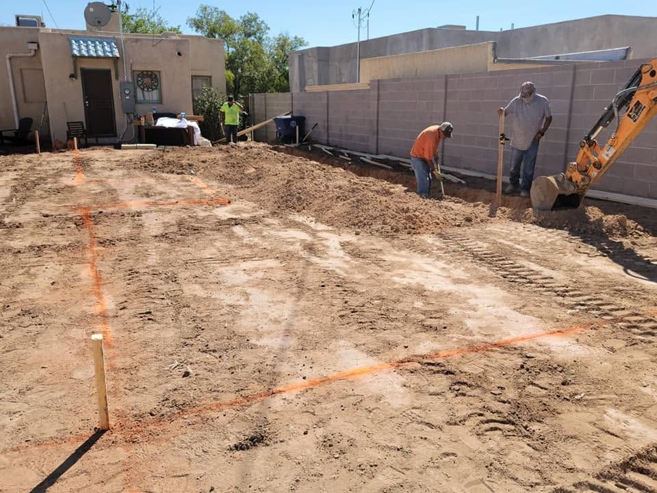 Construction workers grading a dirt lot beside a wall, with an excavator and marked layout lines