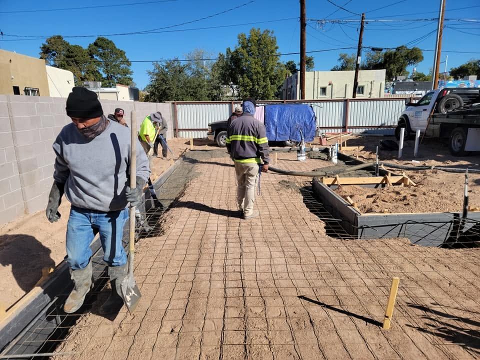 Workers paving a walkway at a construction site with rebar, tools, and a pickup truck nearby.