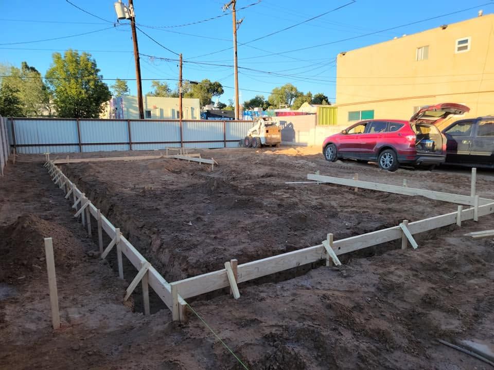 Graded dirt lot bordered by wooden forms and a white fence, with a red SUV parked beside a building.
