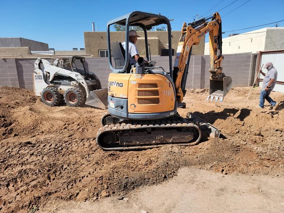 Small excavator and skid steer working on a dirt lot near a building, with a worker standing nearby.