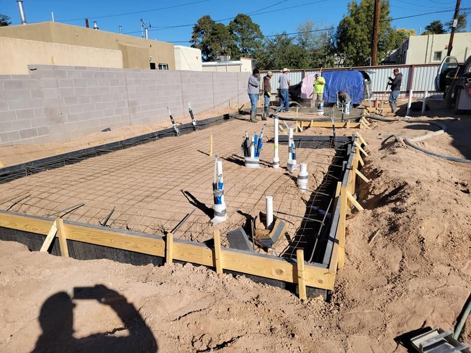 Foundation rebar and plumbing under construction, with workers in the background and wooden forms around the slab