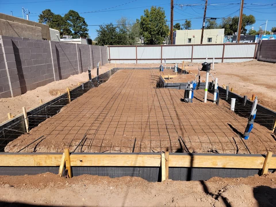 Concrete foundation site with rebar grid, wooden forms, and utility pipes under construction
