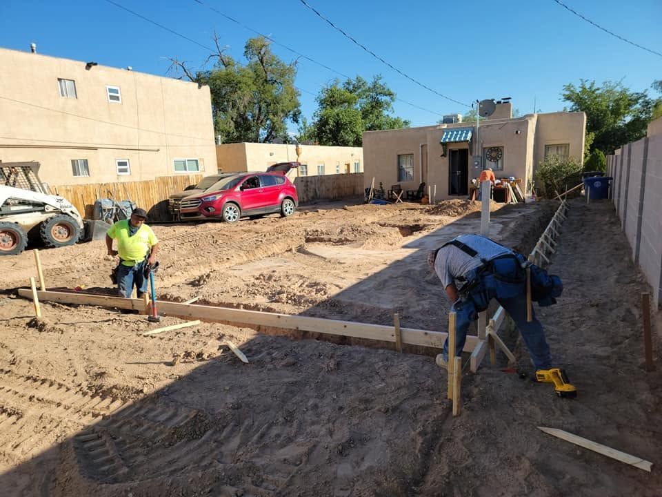 Construction workers leveling dirt in a narrow yard beside beige buildings, with a red SUV parked nearby.