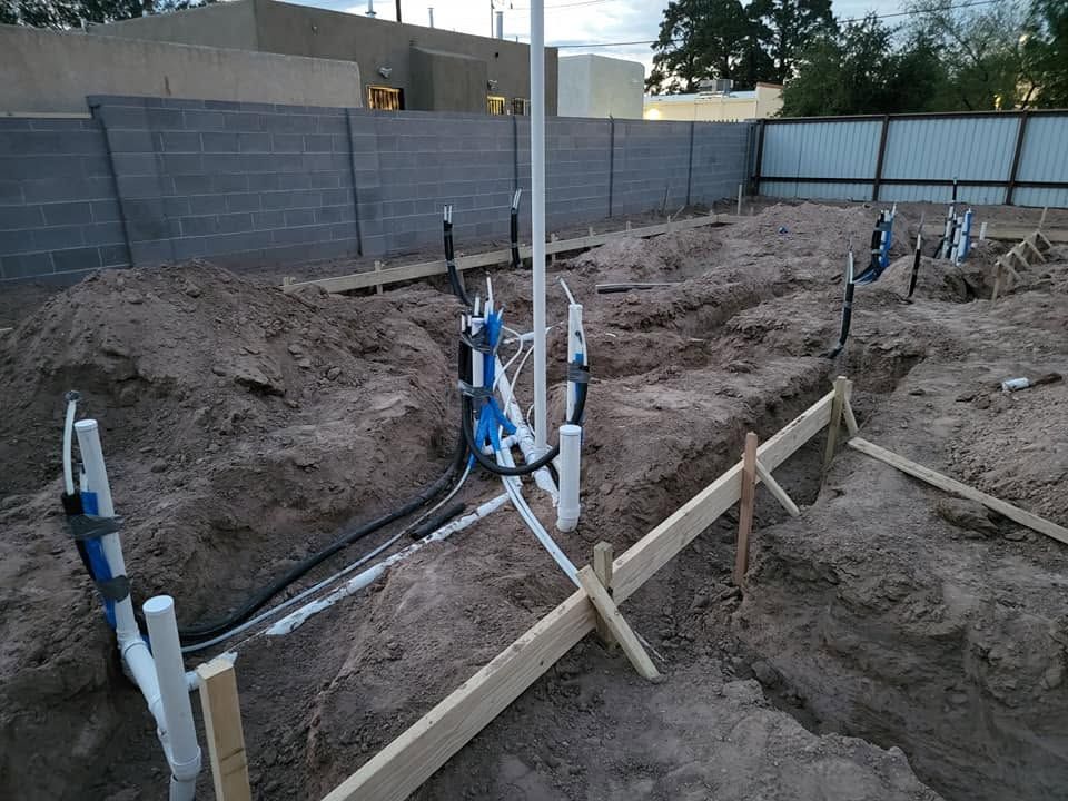 Construction site with trenches, exposed pipes, and wooden stakes in a dirt yard bordered by a gray fence