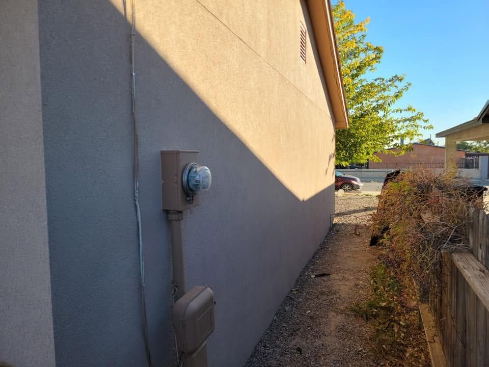 Exterior wall with utility meter and electrical boxes beside a narrow gravel walkway, sunlight casting shadows