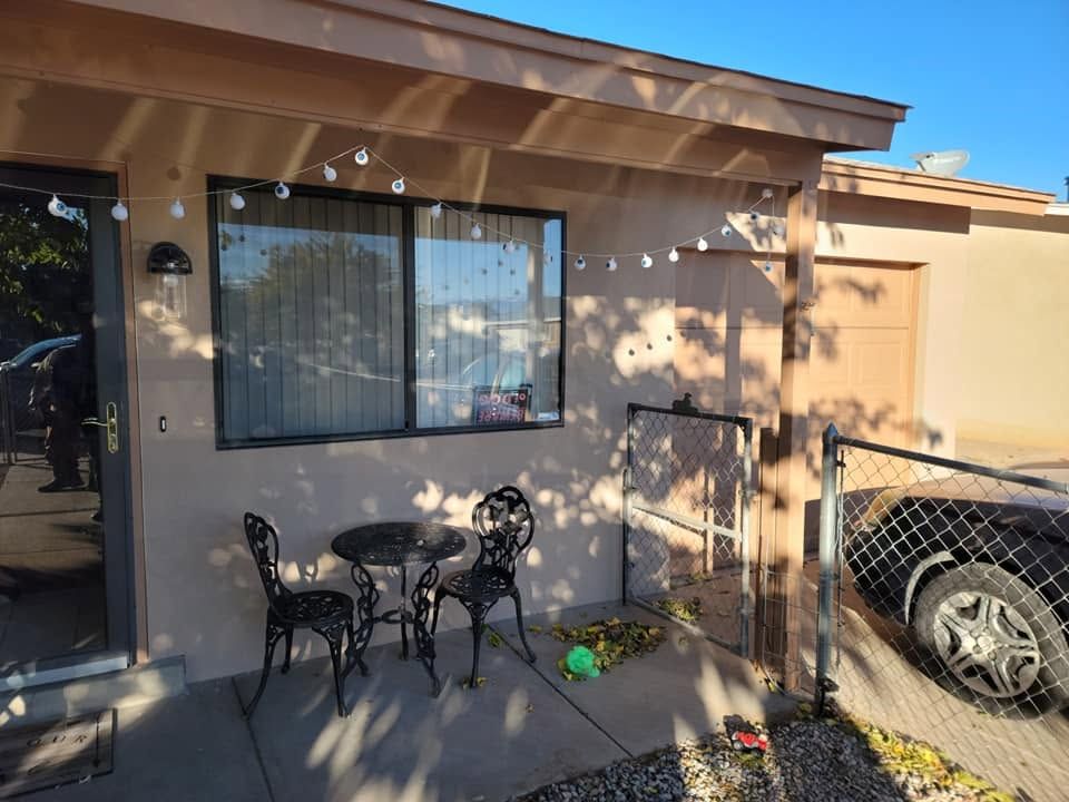 Patio with metal table and chairs beside a beige house, window, potted plants, and a parked car nearby.