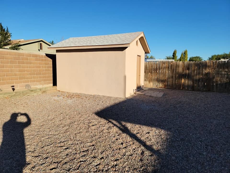 Small beige backyard shed with gravel ground, block wall, and blue sky in a sunny yard.