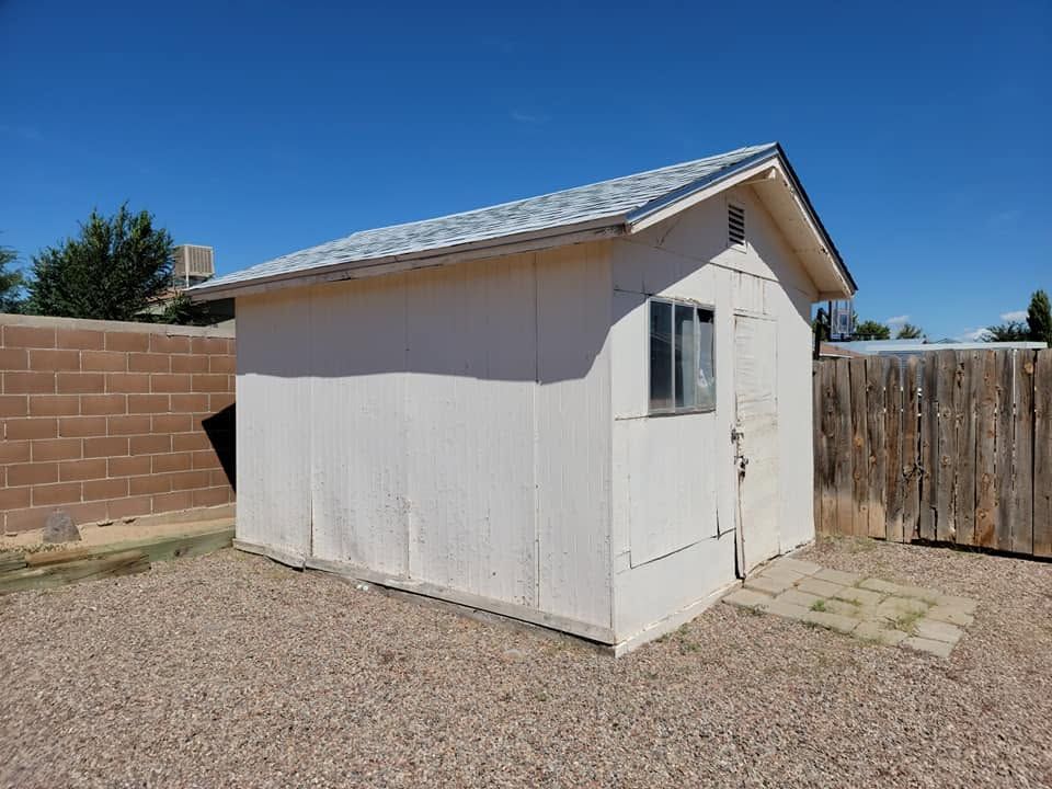 Small white shed with a gray roof in a gravel backyard beside a wooden fence and brick wall