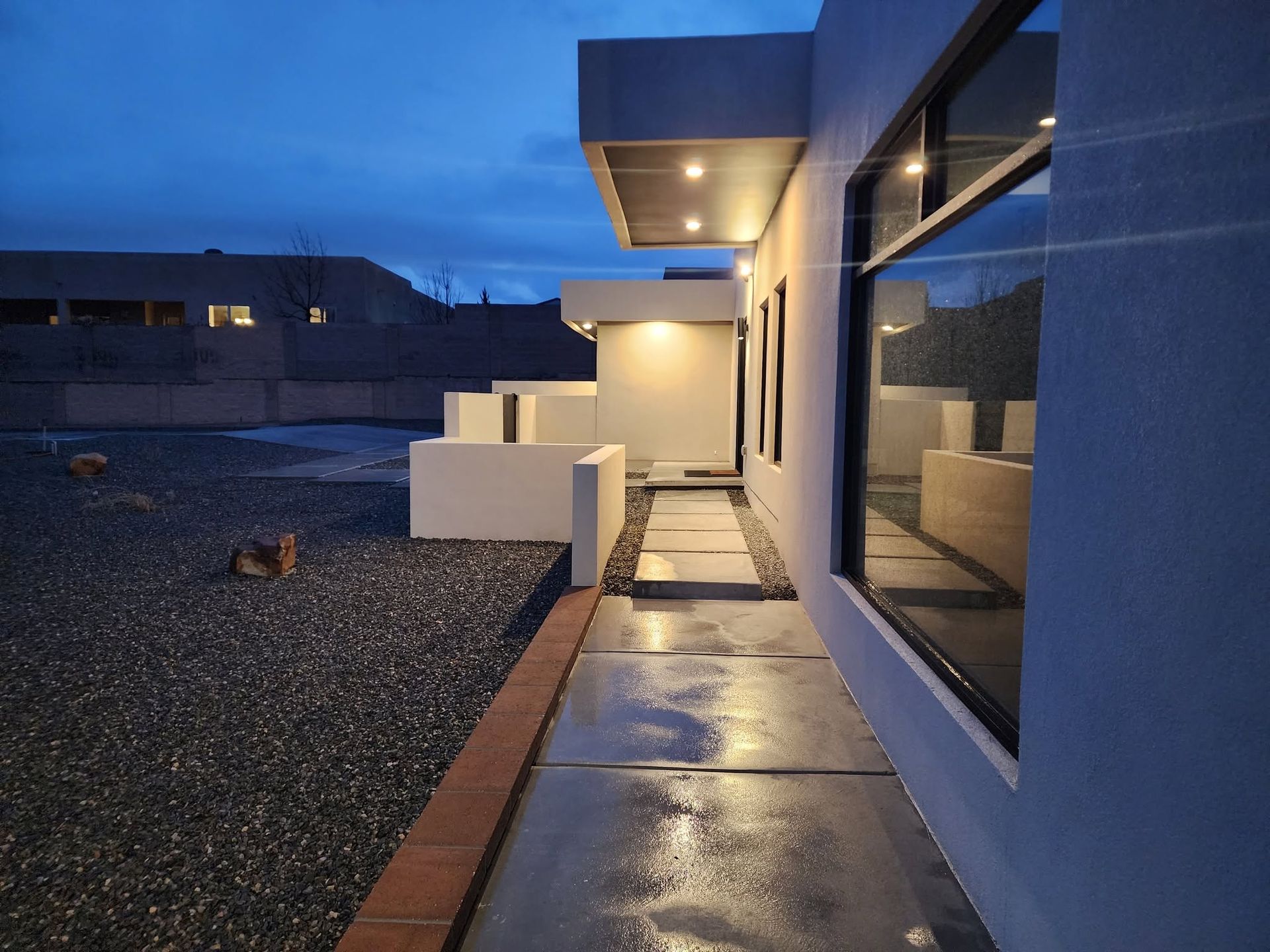 Modern house walkway at dusk with wet concrete, gravel yard, and lit overhangs beside large windows