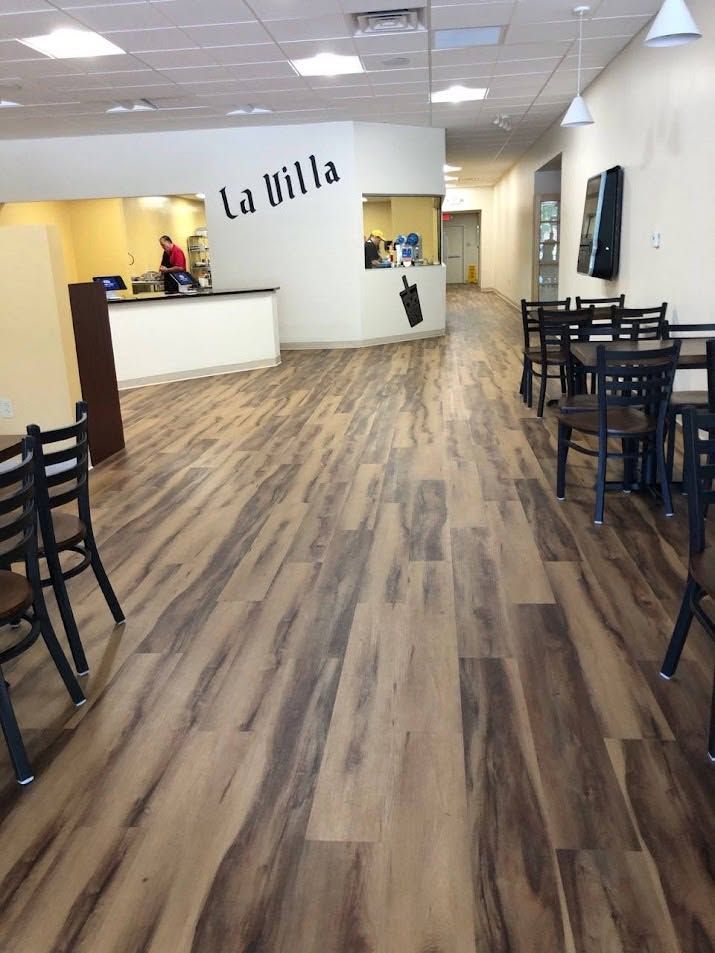 Empty restaurant interior with wood-look floors, tables, and a white front counter labeled “La Italia”