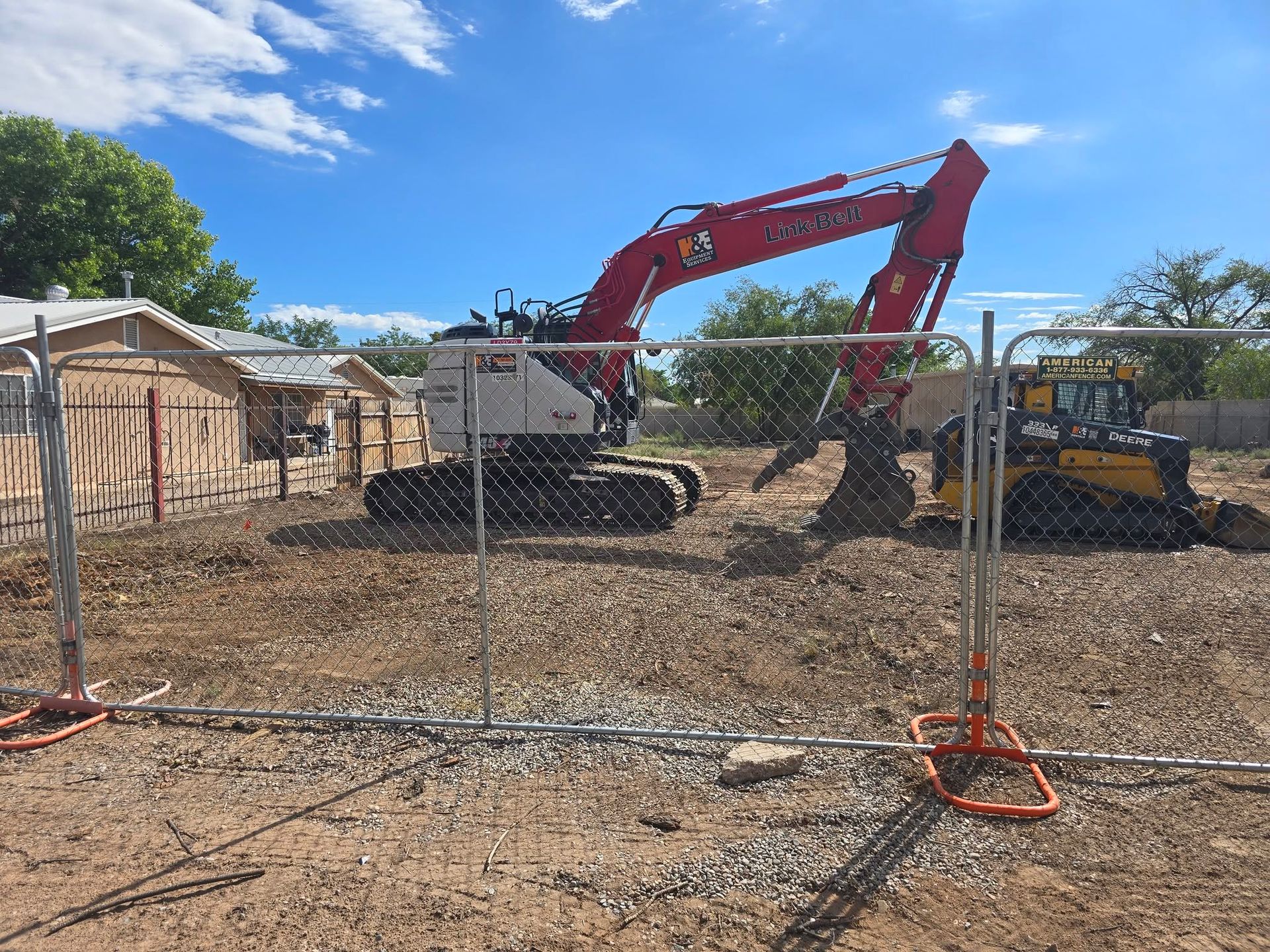 Construction site with red excavator and yellow skid steer behind a wire fence under a blue sky