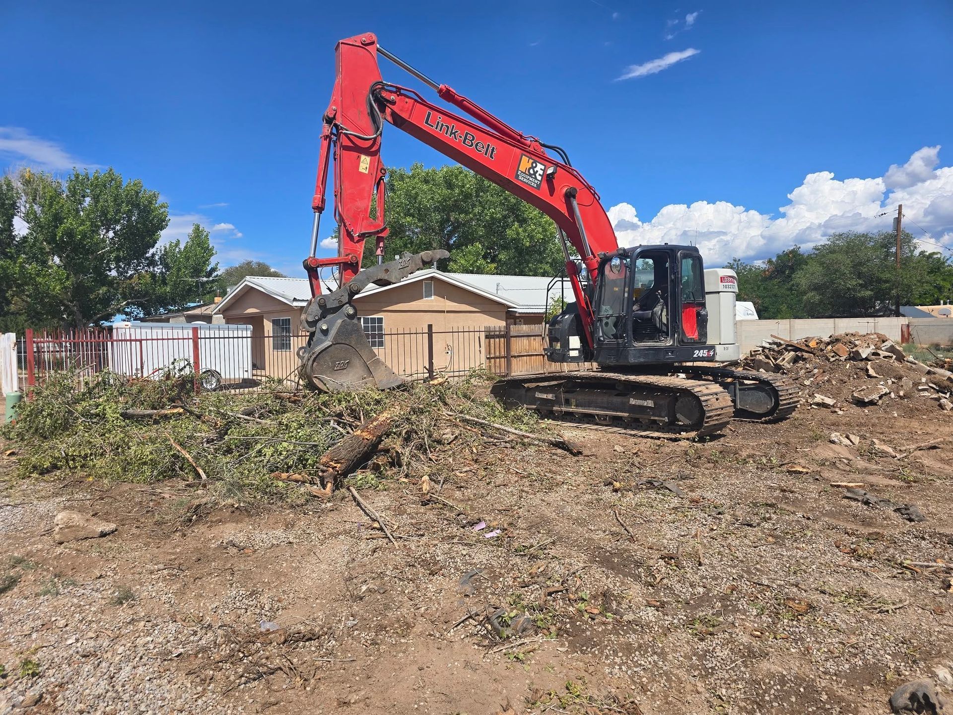 Red excavator clearing dirt on a construction site with houses in the background under a blue sky