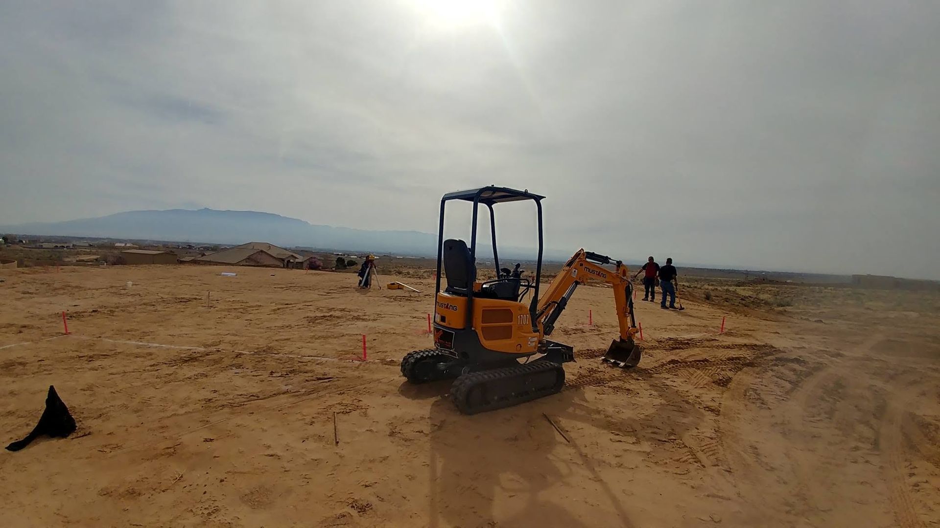 Excavator digging in a sandy construction site under a cloudy sky, with a few people in the distance