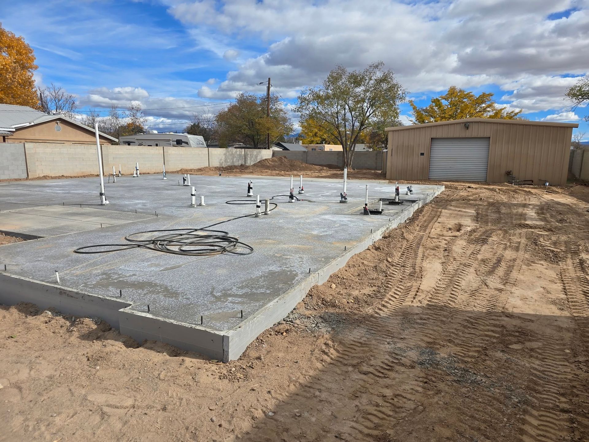 Concrete foundation under construction beside a dirt lot and tan garage under a cloudy sky
