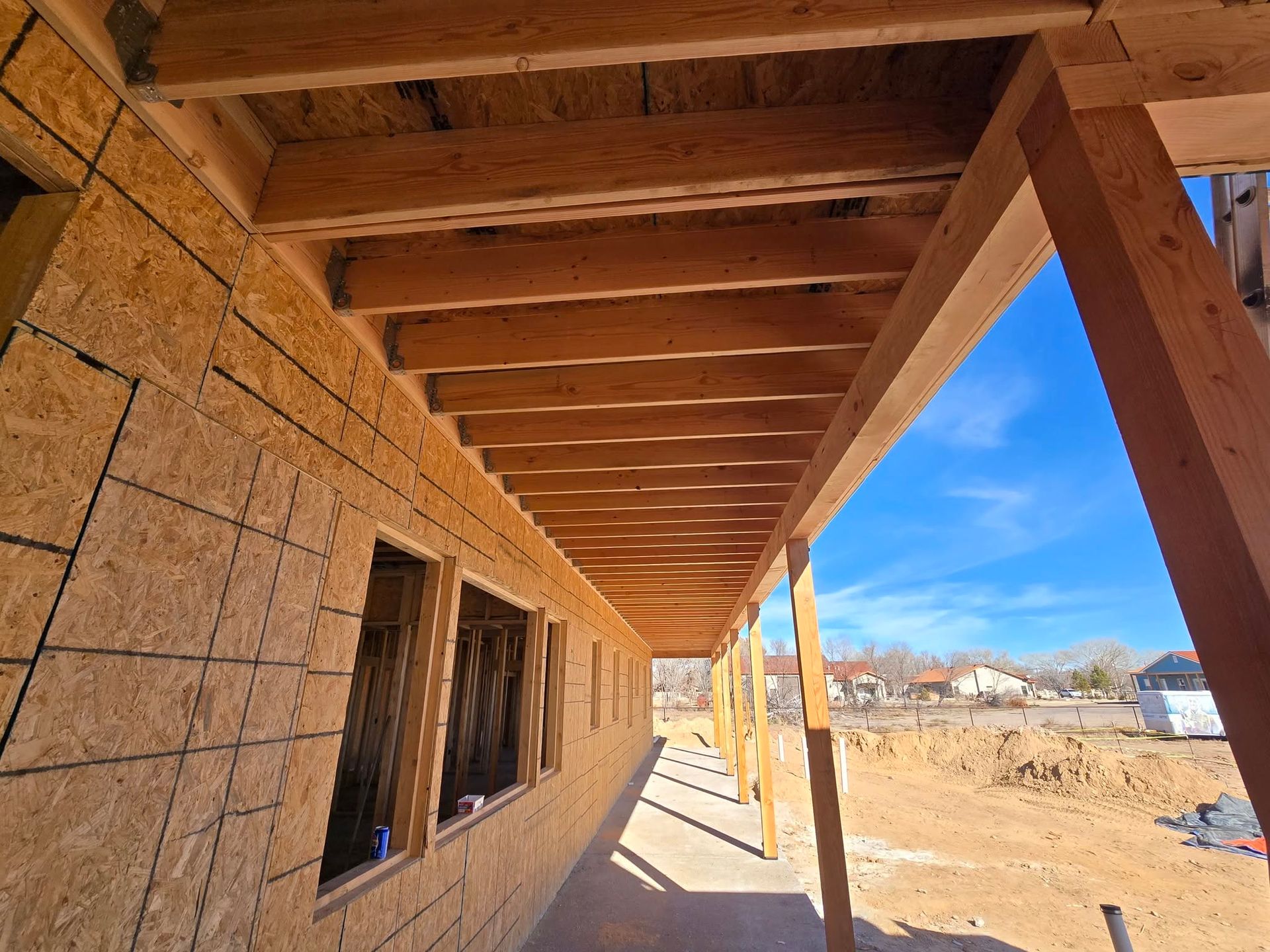 Unfinished house exterior with wooden porch roof and framed windows, looking out onto a sunny dirt lot
