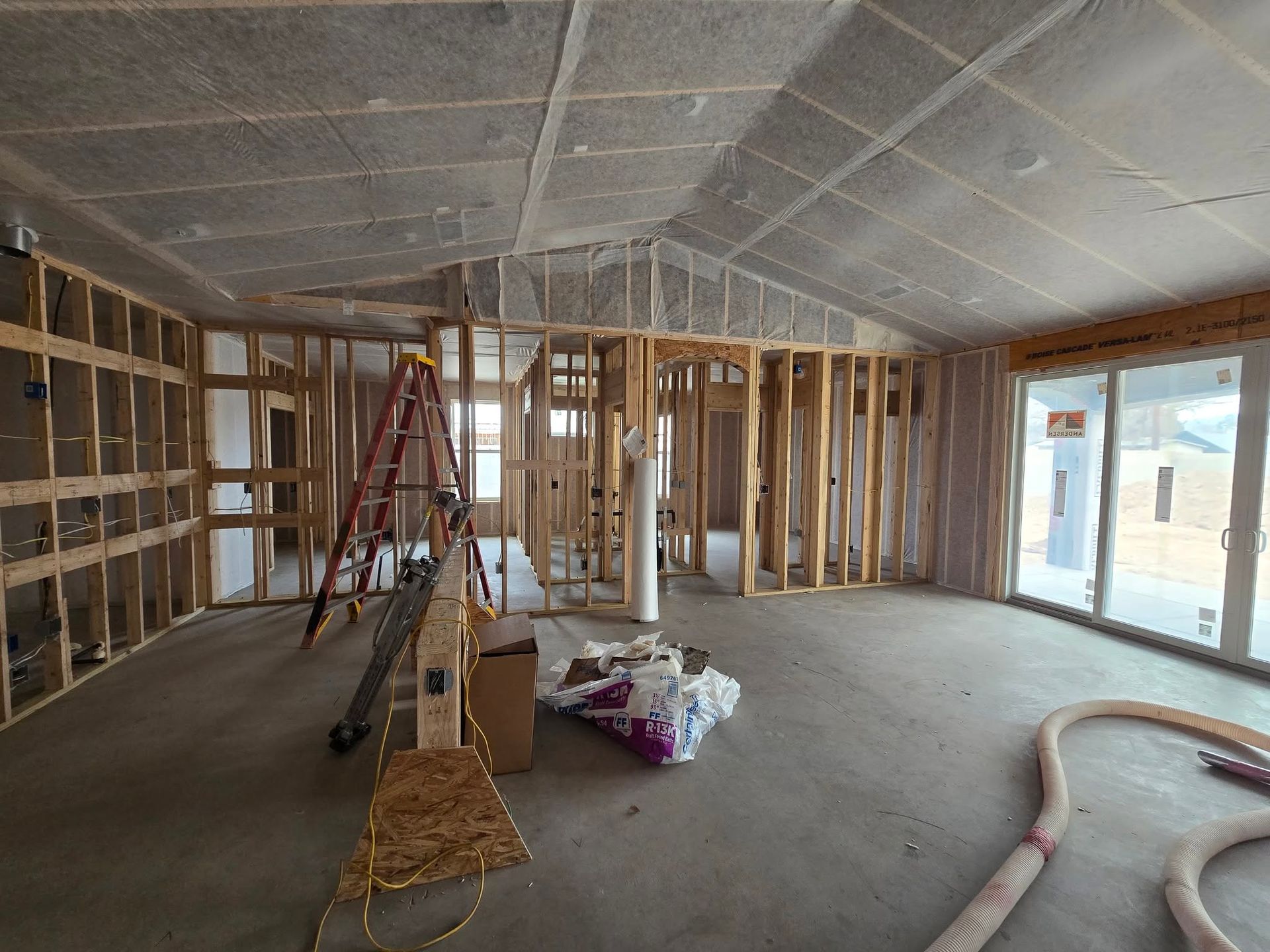 Unfinished room with exposed framing, ladder, construction materials, and large windows along the right wall.