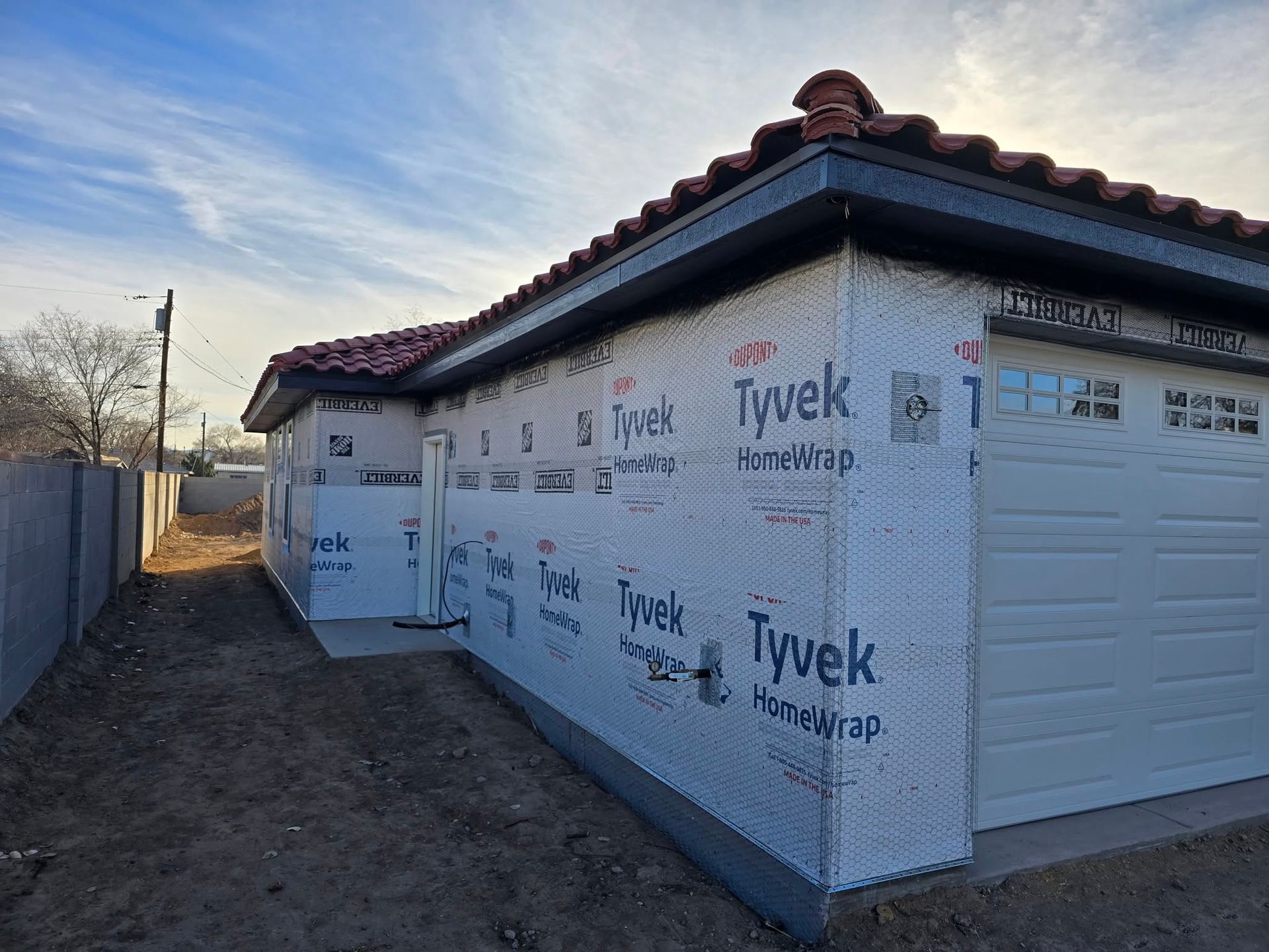 Partially built house with Tyvek wrap, white garage door, and tiled roof in a dirt yard