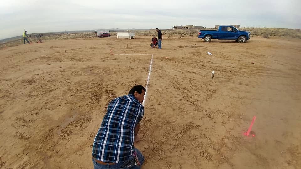 Person crouches on a sandy field, aiming along a white line toward a distant blue pickup truck.