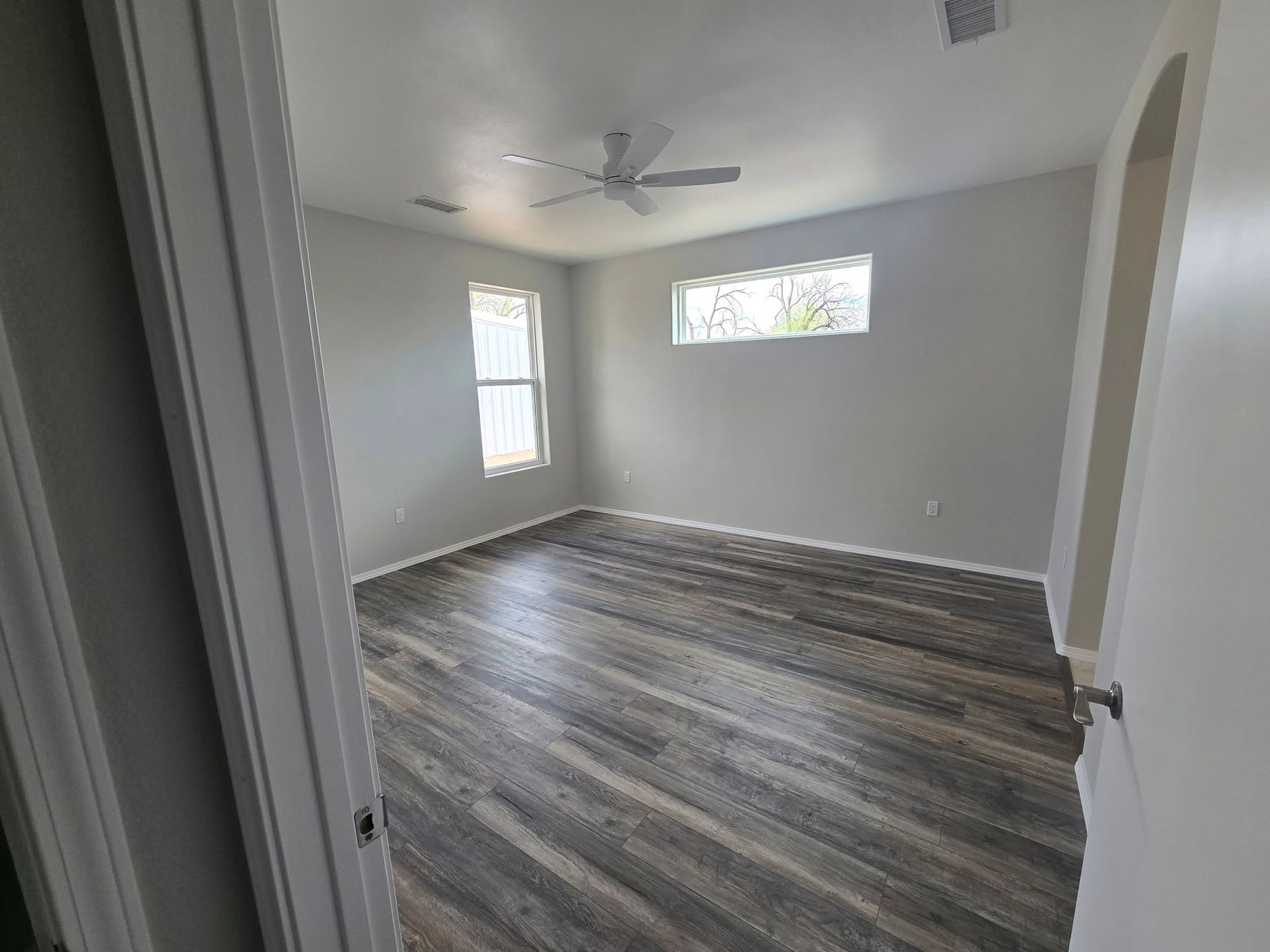 Empty bedroom with gray walls, dark wood-look floors, white ceiling fan, and two windows with blinds