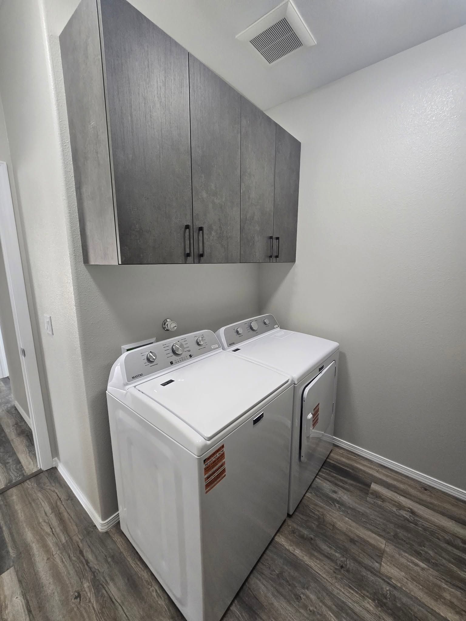 Laundry room with white washer and dryer beneath dark gray cabinets on wood-look flooring