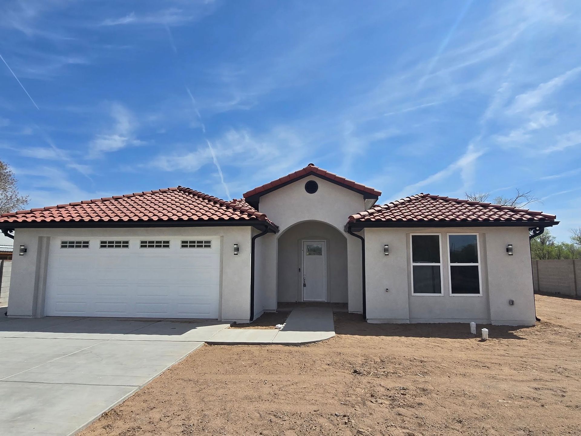 Single-story stucco house with a red tile roof, two-car garage, and dry front yard under a blue sky