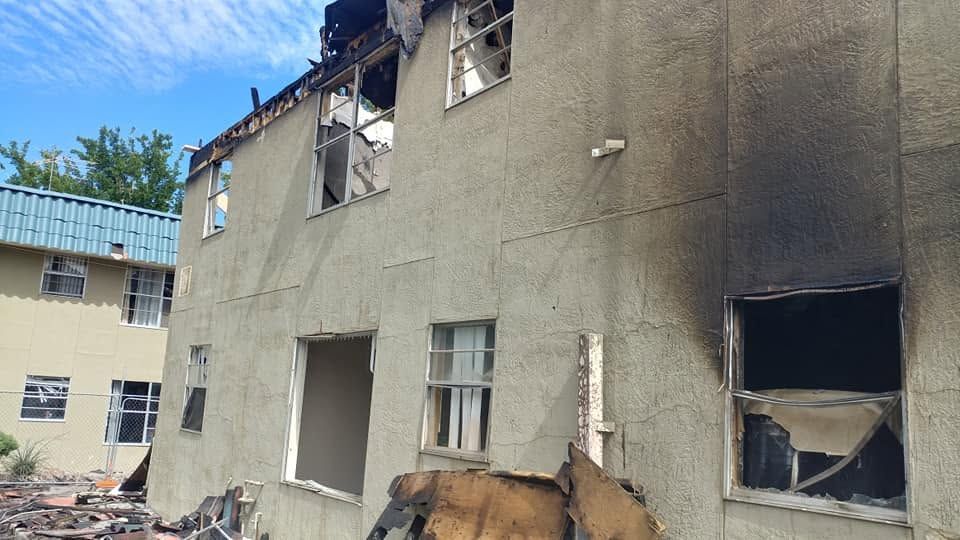 Damaged two-story building with shattered windows, cracked walls, and debris under a blue sky