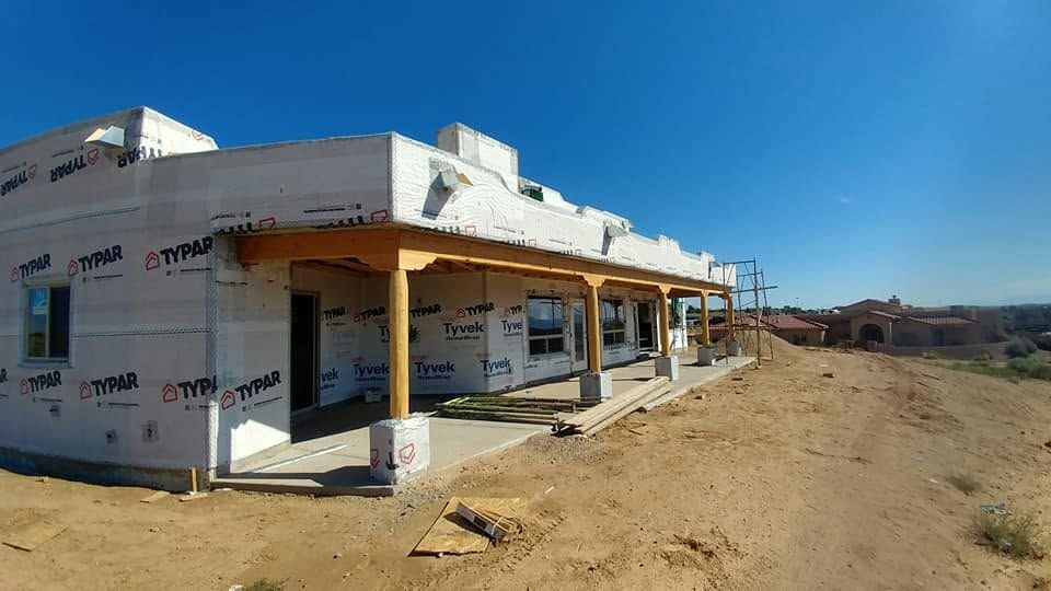 Partially built house with wraparound porch on a dusty construction site under a clear blue sky