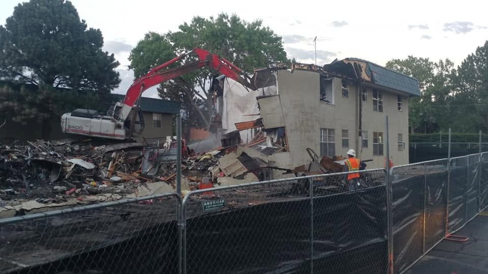 Demolition site with excavator tearing down a damaged building behind a chain-link fence.