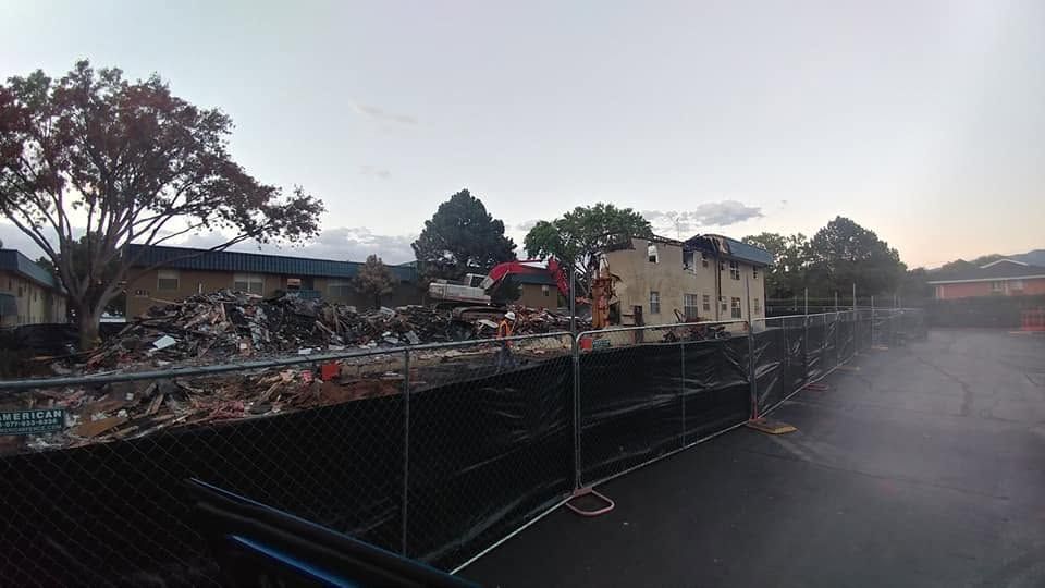 Collapsed building with debris behind a fence at dusk, with trees and smoke or dust in the foreground
