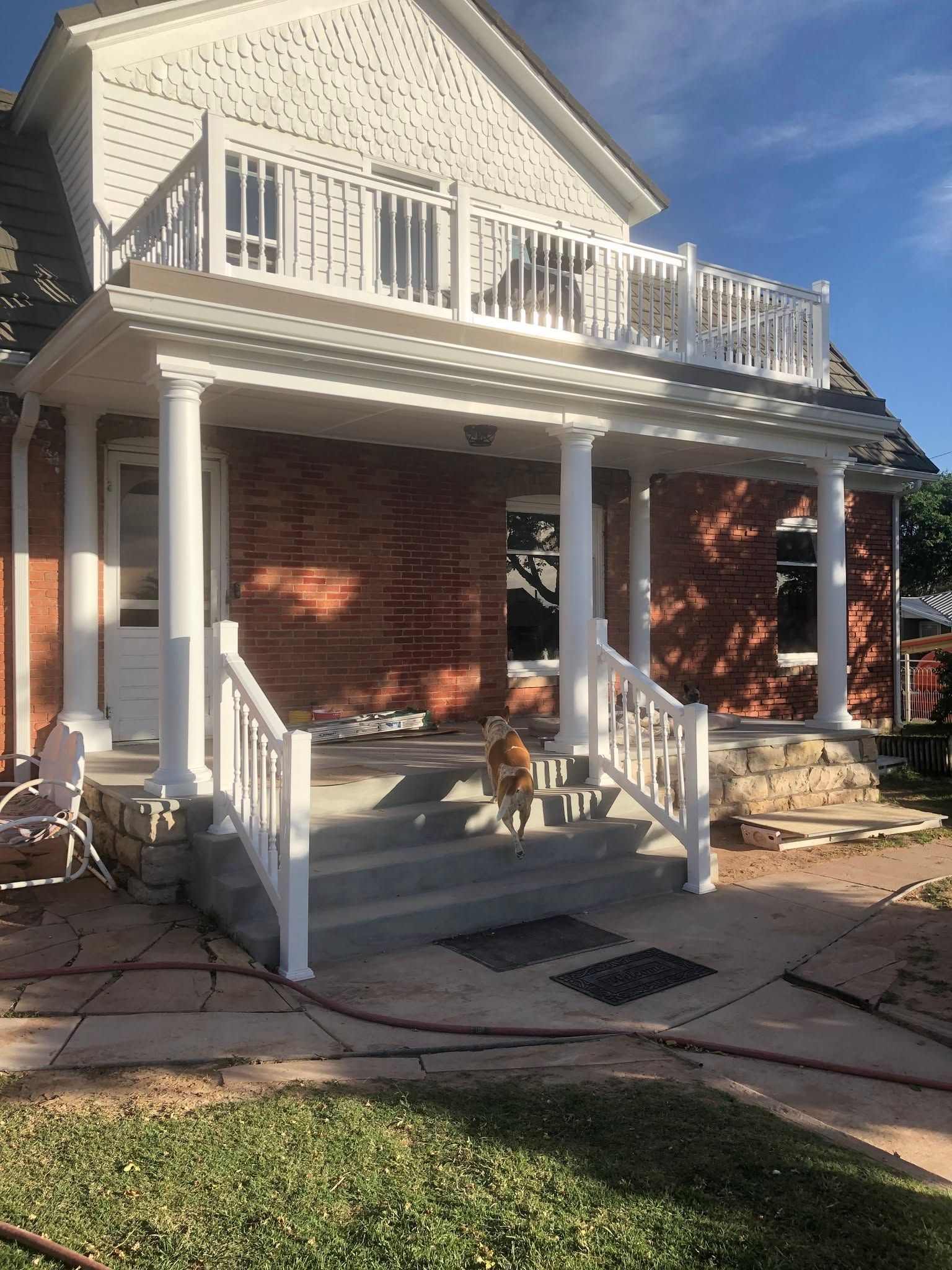 Front porch of a red brick house with white railings, columns, and a balcony in bright sunlight