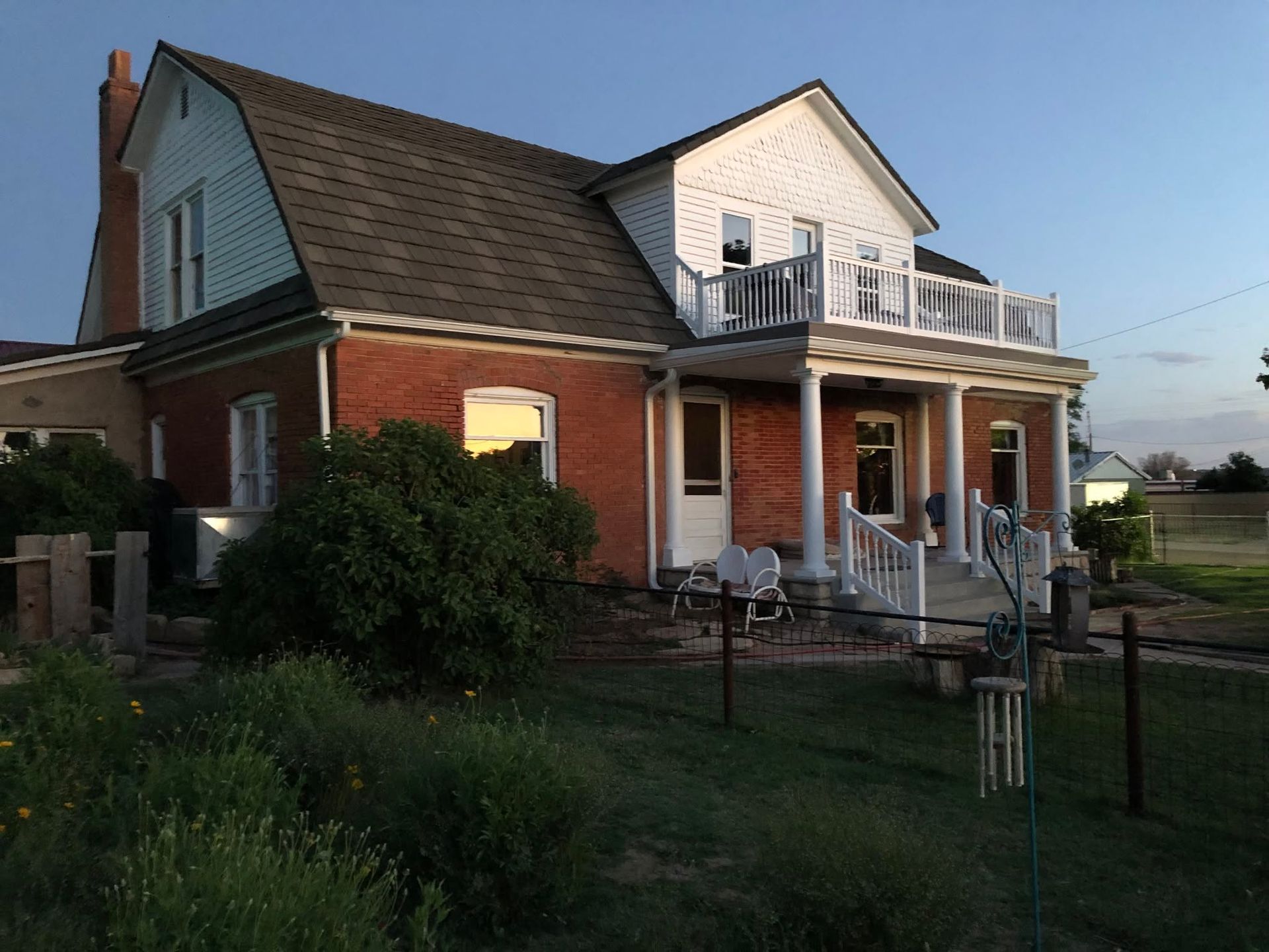Brick house with white upper story, front porch, and balcony in a grassy yard at dusk