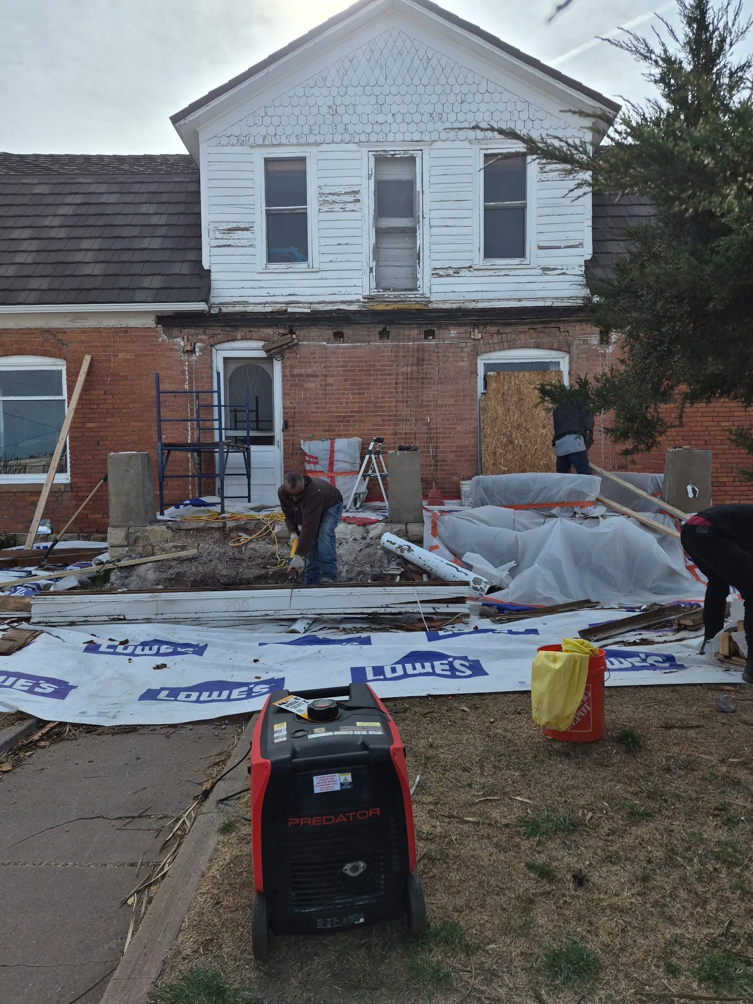 Damaged brick house with torn roof tarps, debris in the yard, and construction materials outside.