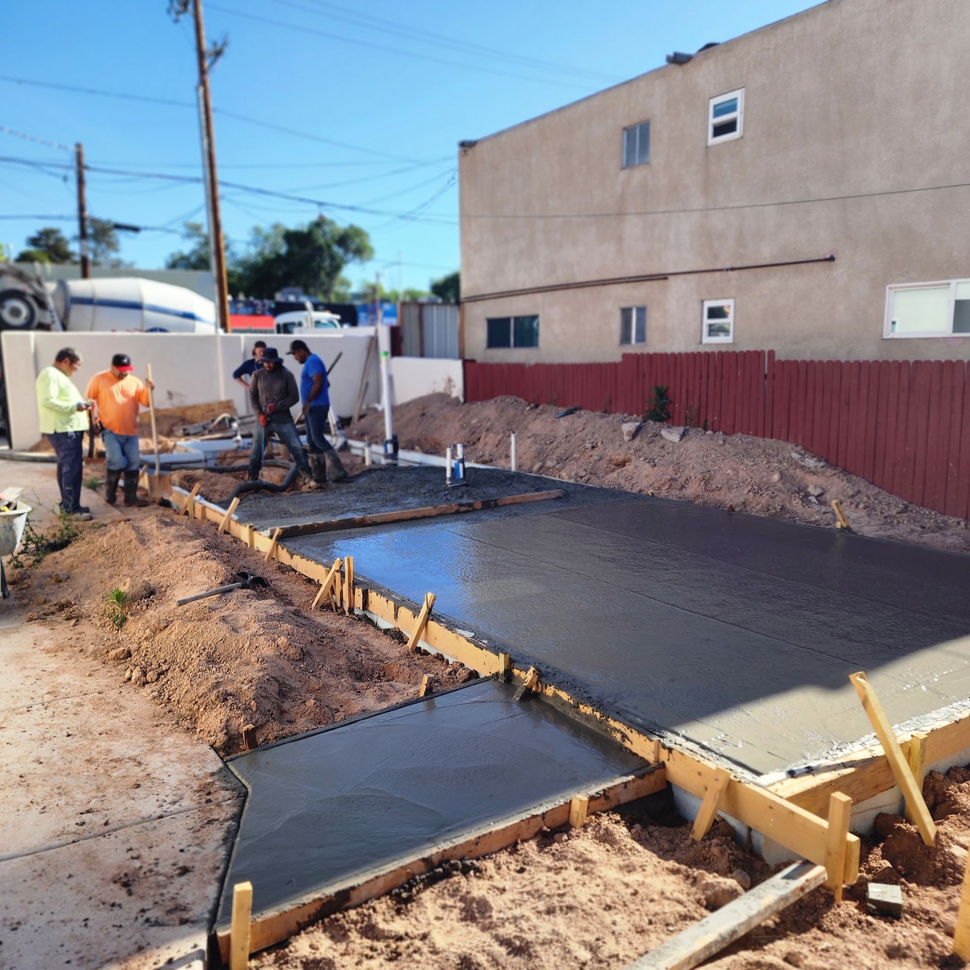 Workers pouring fresh concrete for a sidewalk beside a building on a sunny street.