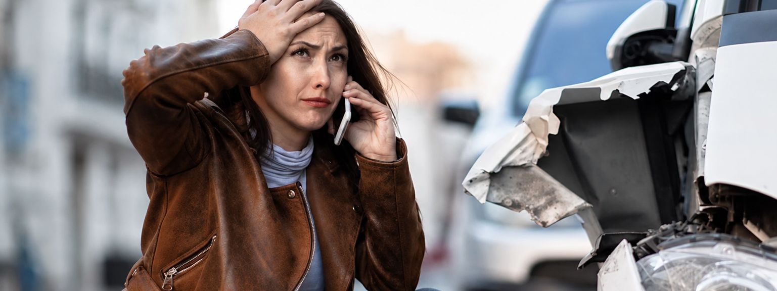 Woman in a brown coat looking stressed beside a damaged car on a snowy roadside.