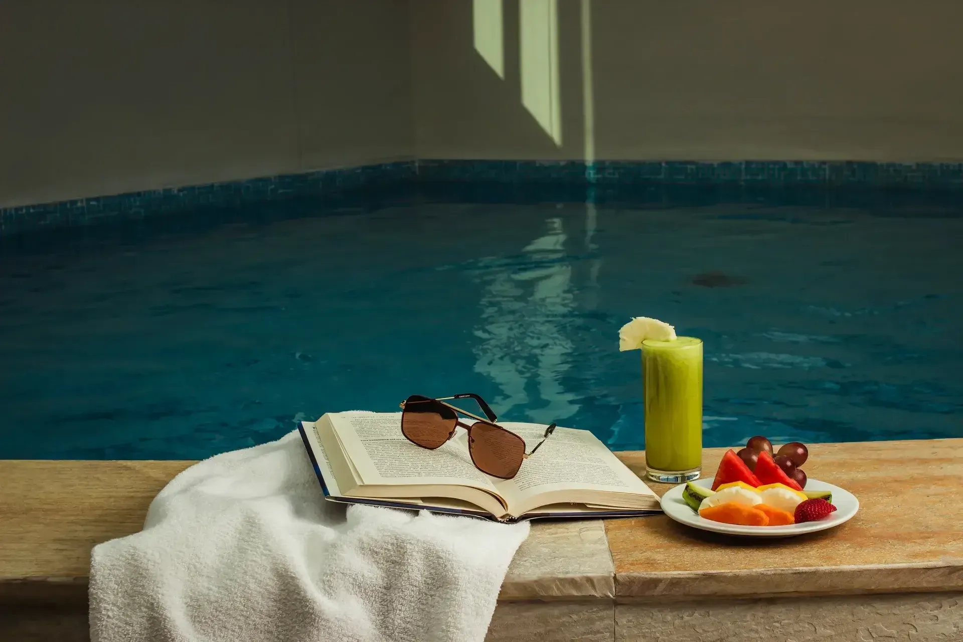 um livro, óculos escuros, um prato de frutas e uma bebida estão à beira de uma piscina.
