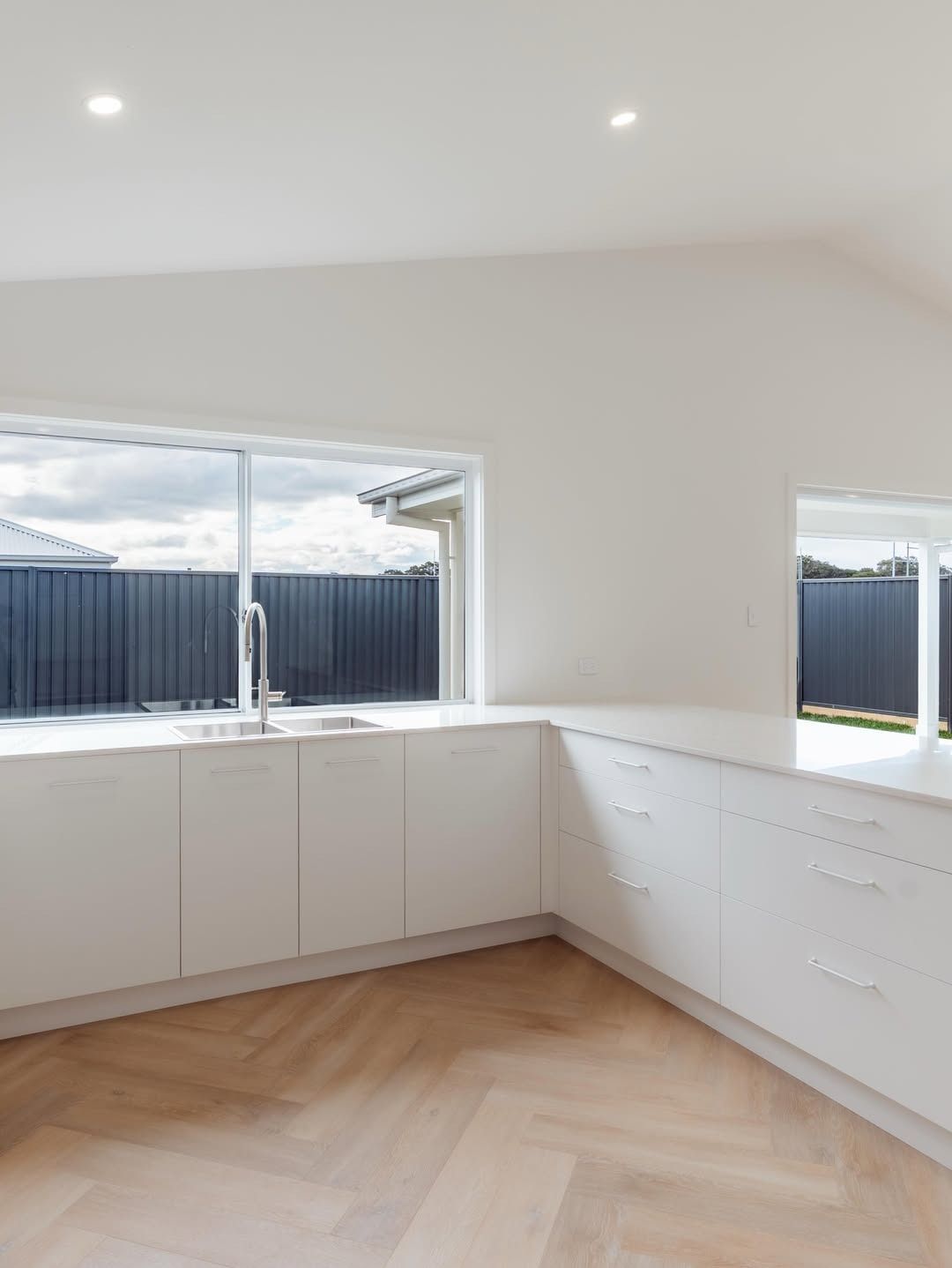 Bright White Kitchen with Corner Cabinets, Herringbone Wood Floor, and Large Windows Showing a Fenced Yard — JDS Kitchens & Detailed Joinery in Albion Park Rail, NSW
