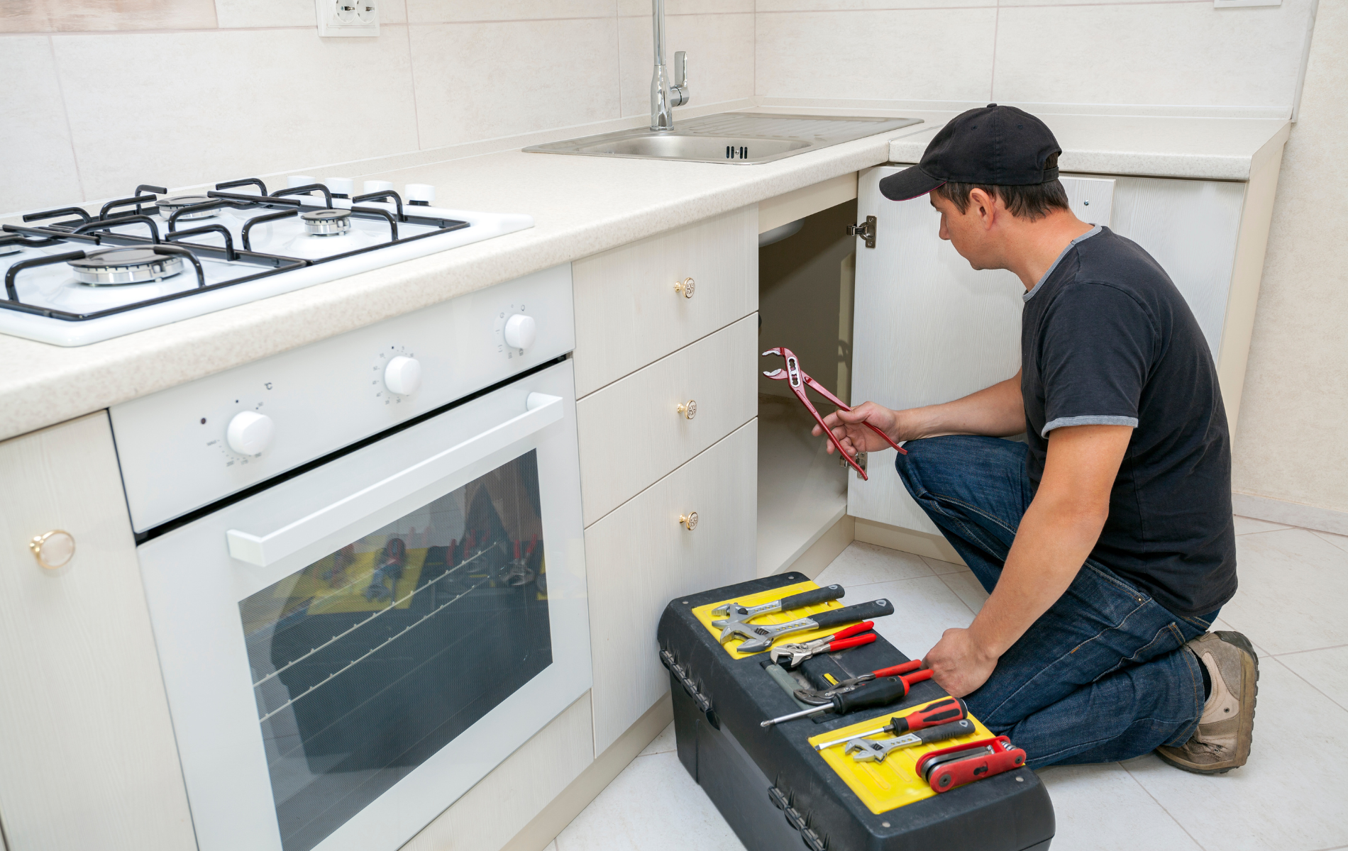 A man is kneeling down in a kitchen with a toolbox.
