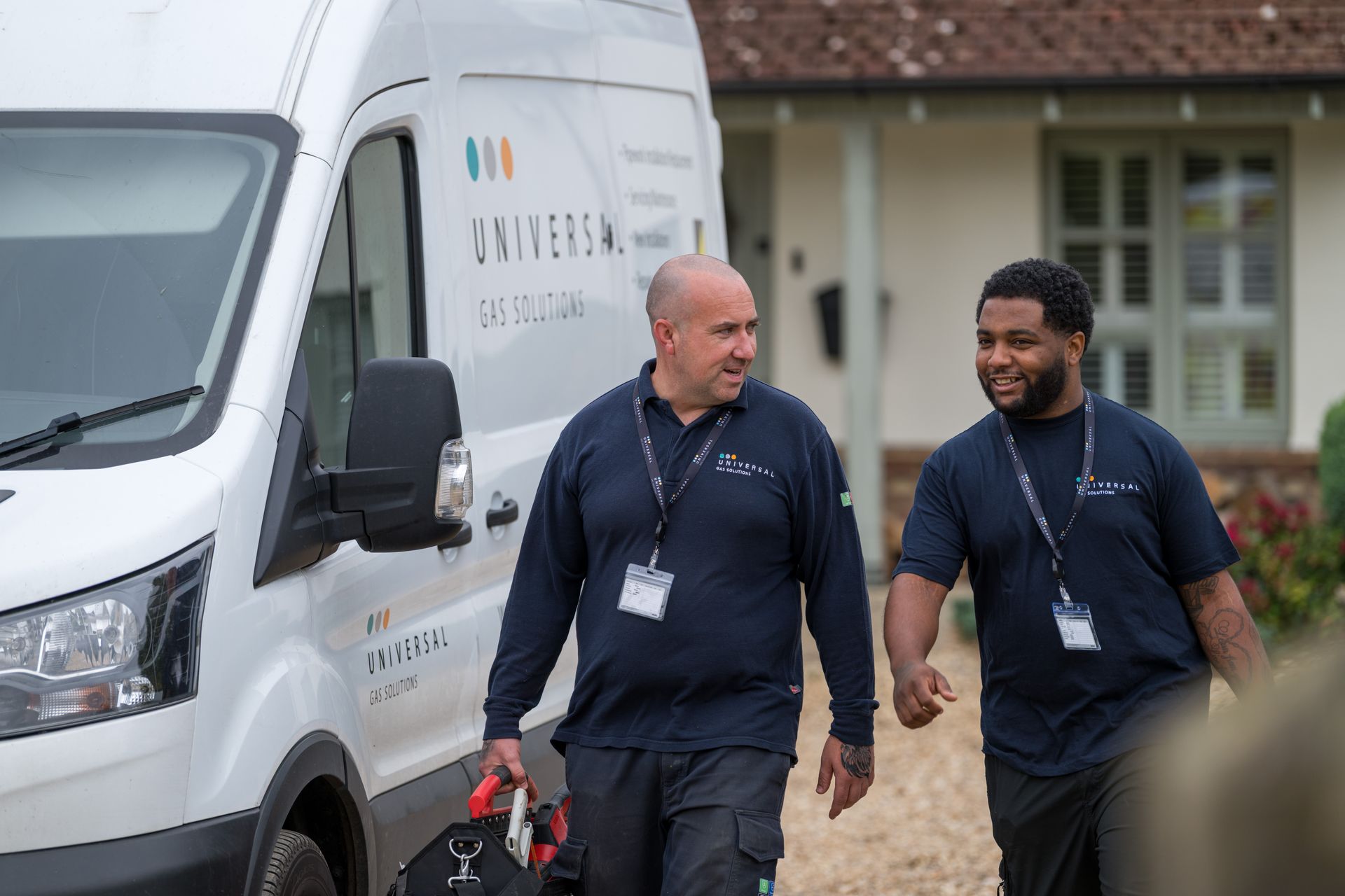 Two men in navy uniforms walk near a van with a logo. One carries a tool bag.