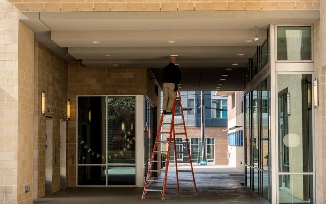 Person on a ladder repairing the ceiling of a building entrance with glass doors.
