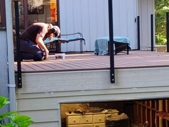 Person kneeling on a deck, working with tools near a covered chair by a house wall.
