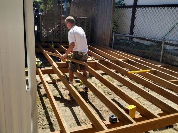 Man installing wooden deck framing in a backyard beside a house and fence